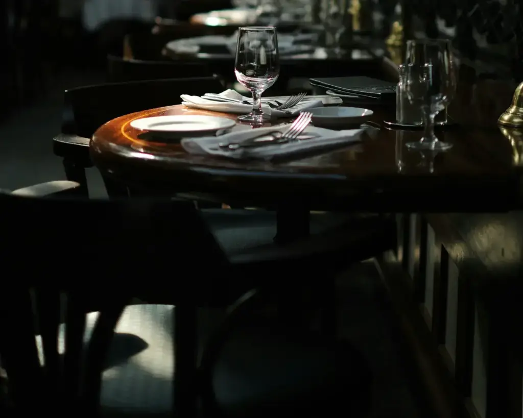 Dimly lit restaurant table set with elegant glassware, plates, and cutlery. The warm light casts soft shadows, creating an intimate, cozy atmosphere.