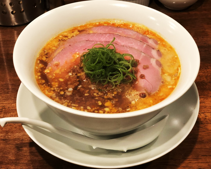 A bowl of ramen with a rich broth, topped with several slices of pink char siu pork and garnished with a heap of green onions, on a wooden table.