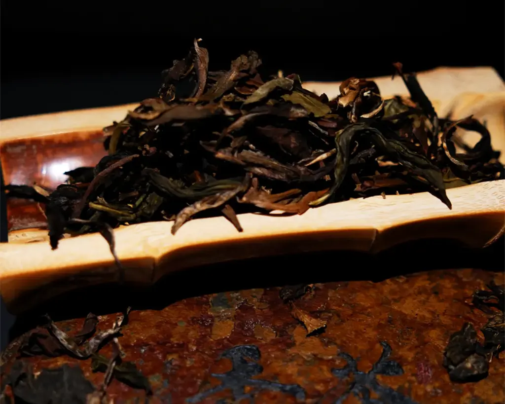 Dried tea leaves rest on a wooden tray against a dark background, conveying a rustic and earthy feel. The textures of leaves and wood are prominent.