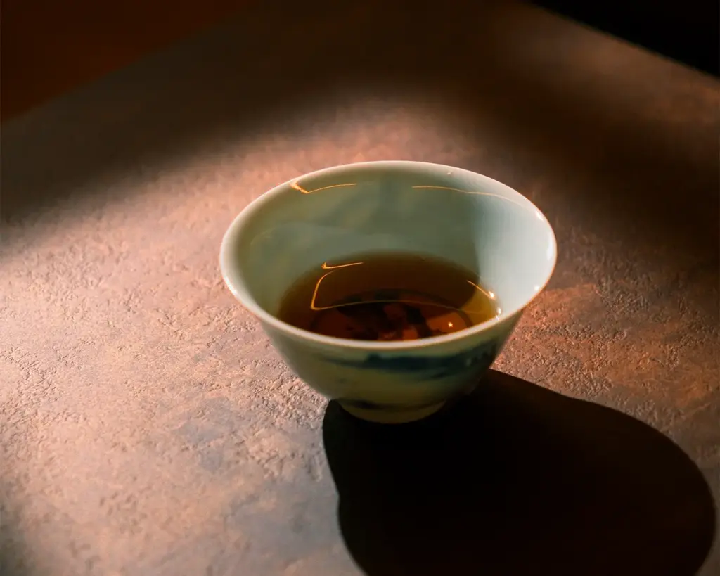 A small porcelain teacup filled with tea is lit warmly from the side, casting a soft shadow on a textured, earthy surface, evoking a serene and cozy mood.