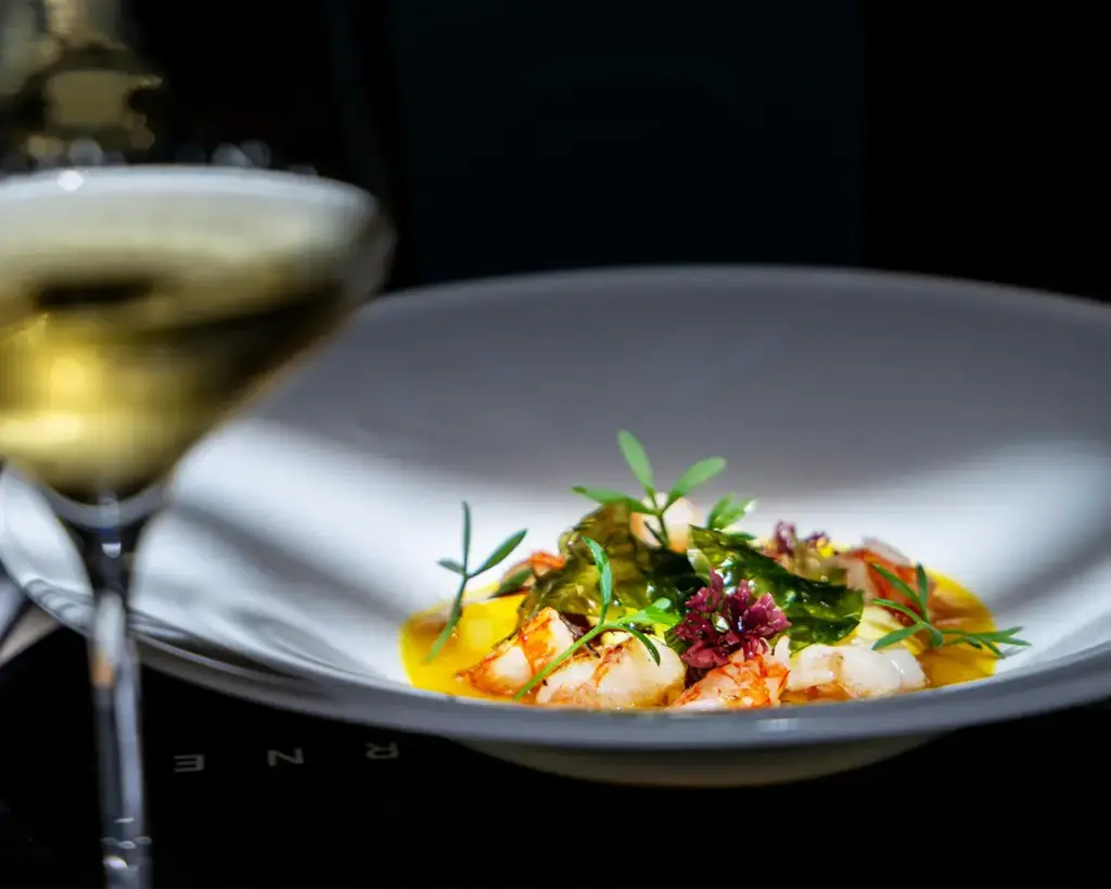 A gourmet dish elegantly presented in a white bowl, featuring shrimp, vibrant green herbs, and a rich sauce. A glass of white wine is blurred in the foreground.