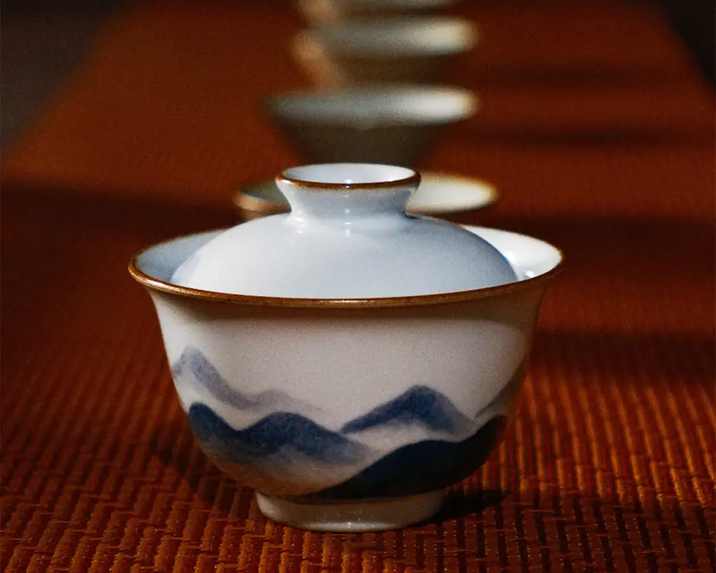 A porcelain teacup with a lid, adorned with blue mountains, sits on a textured orange surface. Three blurred cups line up behind, creating depth.
