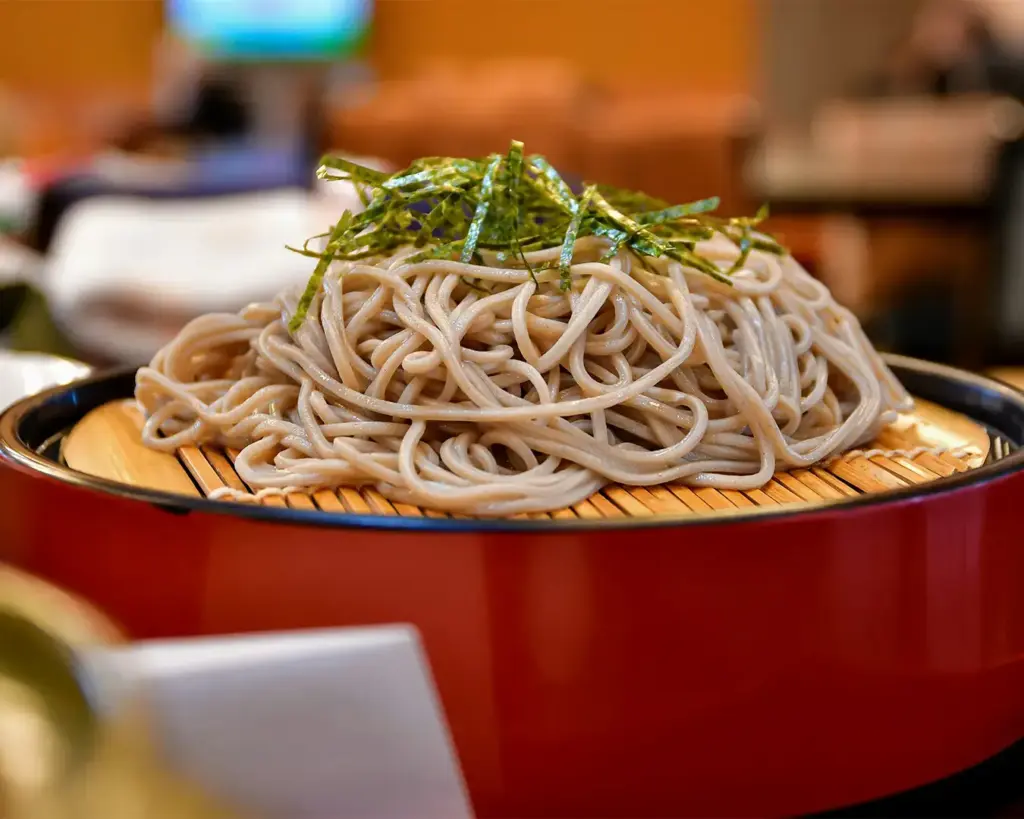 A plate of chilled soba noodles topped with sliced nori is served on a bamboo mat in a red bowl, creating a fresh and inviting presentation.