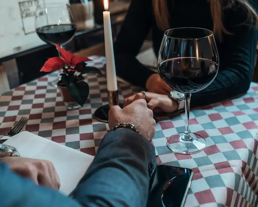 A couple holds hands across a candlelit table in a cozy restaurant. Red wine glasses and a small plant add to the intimate, romantic setting.