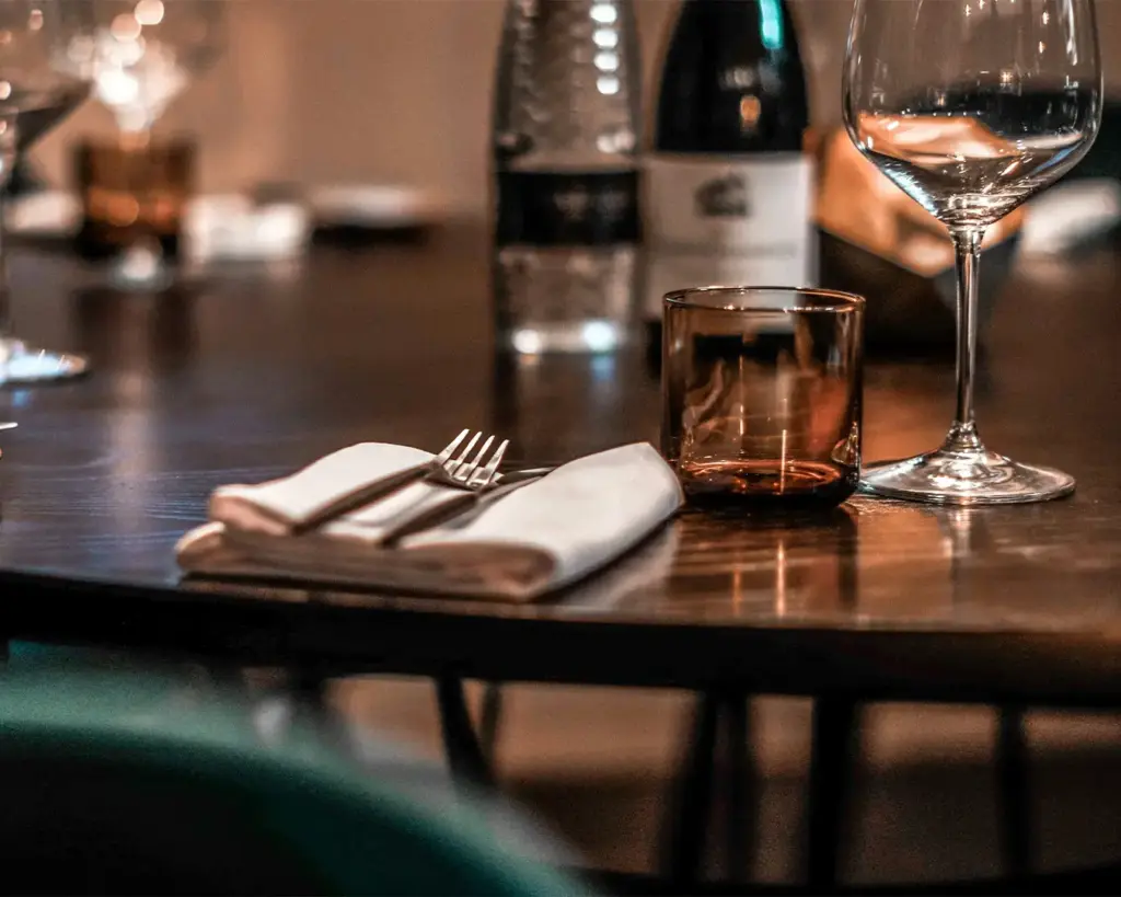 Elegant restaurant table setting with a folded napkin and silverware beside an empty brown tumbler and wine glass, exuding a cozy, intimate ambiance.