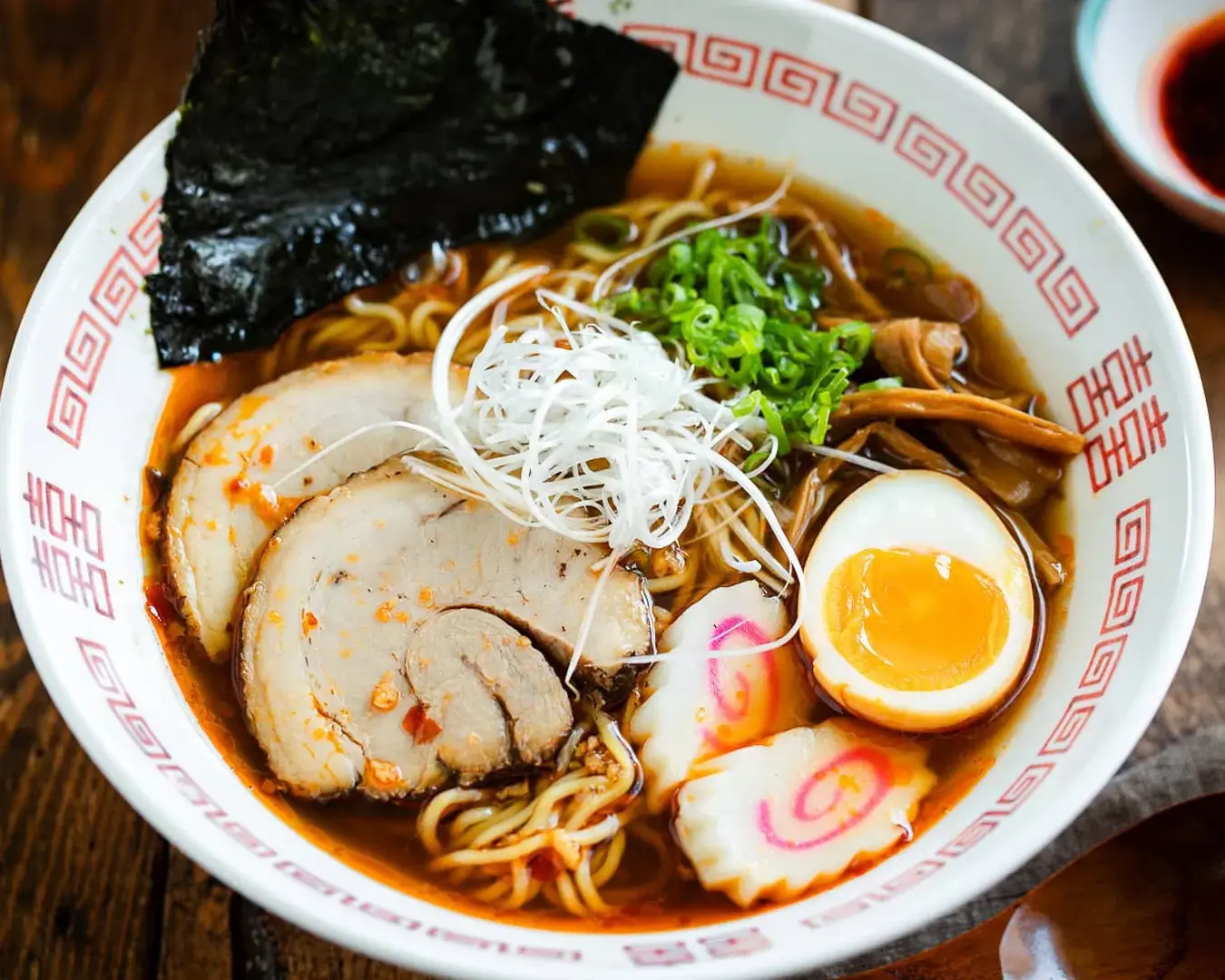 A steaming bowl of ramen with broth, topped with sliced pork, half a soft-boiled egg, green onions, seaweed, and narutomaki, creating a warm, savory appeal.
