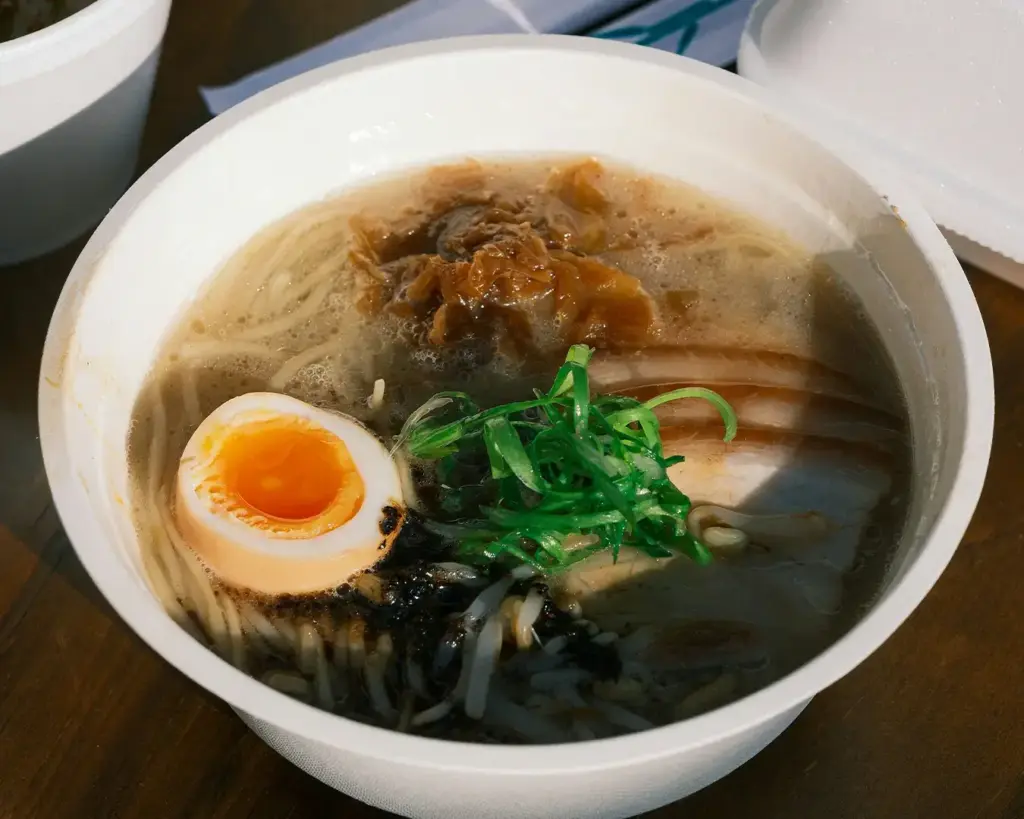Bowl of ramen featuring sliced pork, half a soft-boiled egg, green onions, and a rich broth. The setting is warm and inviting.