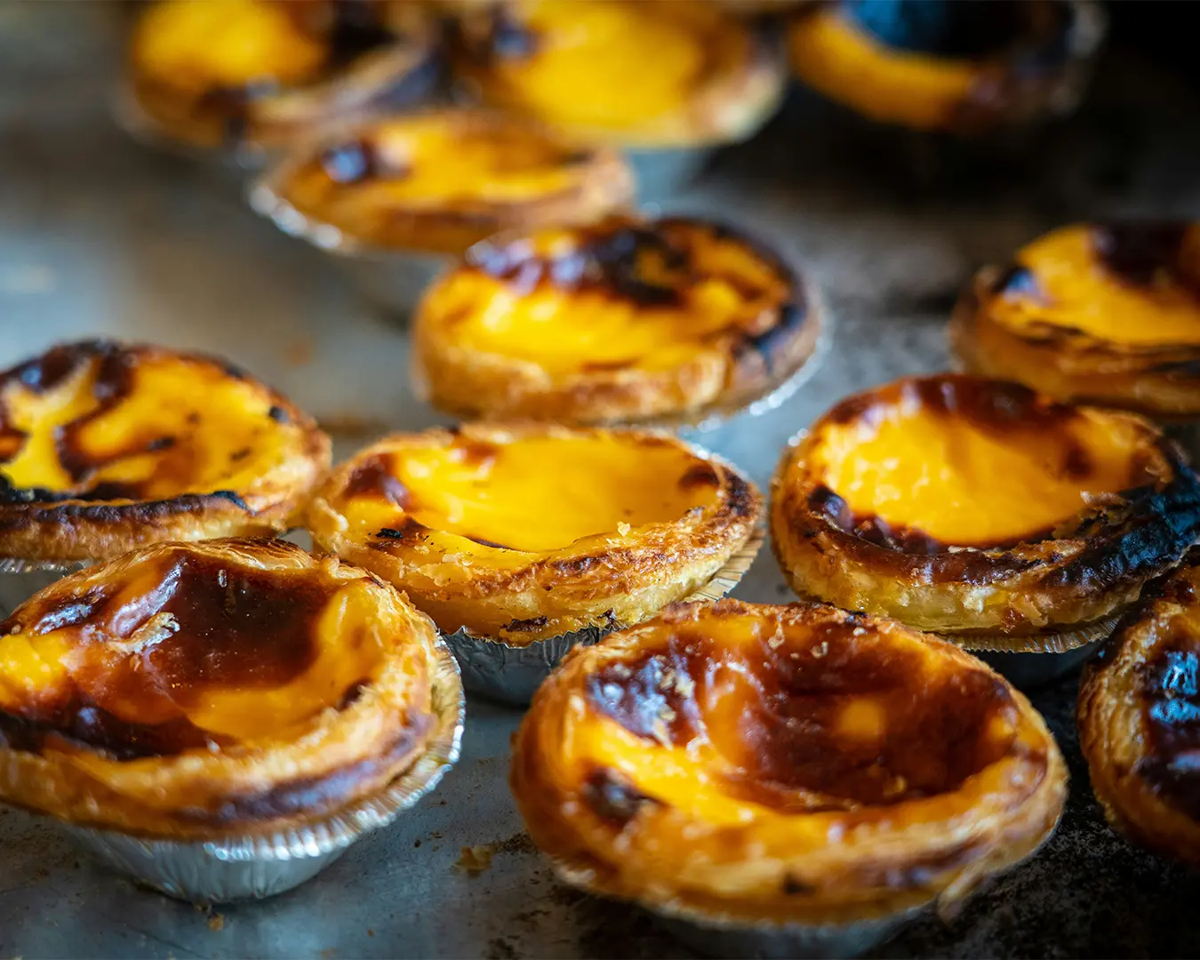 Golden-brown custard tarts with flaky crusts sit on a baking tray. The tarts are slightly caramelized, evoking a warm, inviting atmosphere.