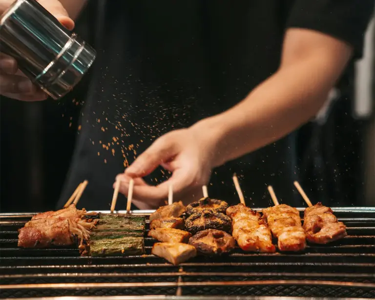 Hands seasoning skewers of grilled meat and vegetables with spice. The skewers are evenly placed on a hot grill, creating a savory, appetizing scene.