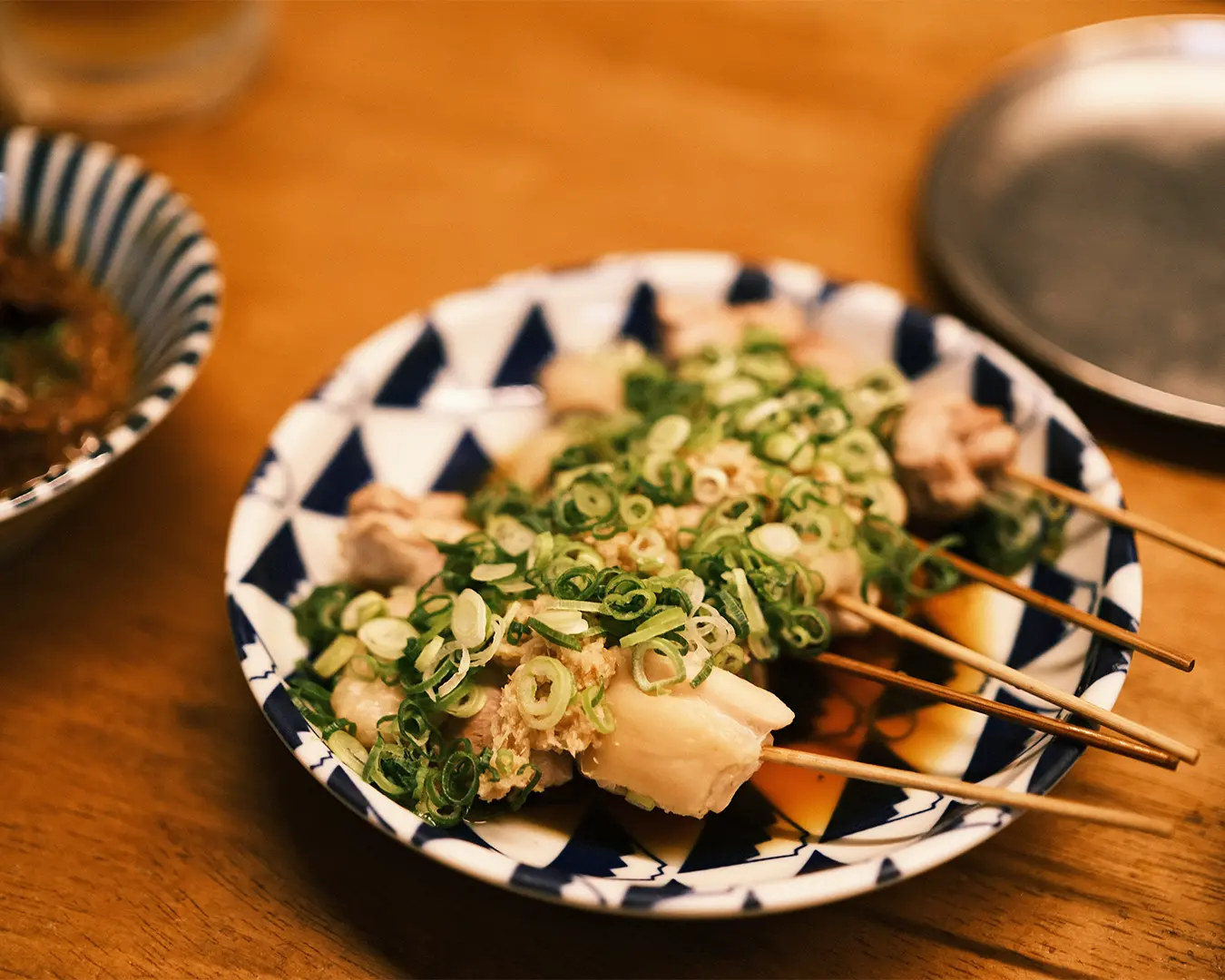 A close-up shot of Japanese yakitori chicken skewers served on a traditional blue and white patterned ceramic plate, generously topped with freshly chopped green onions and a savory tare soy sauce glaze in a Singapore restaurant setting.
