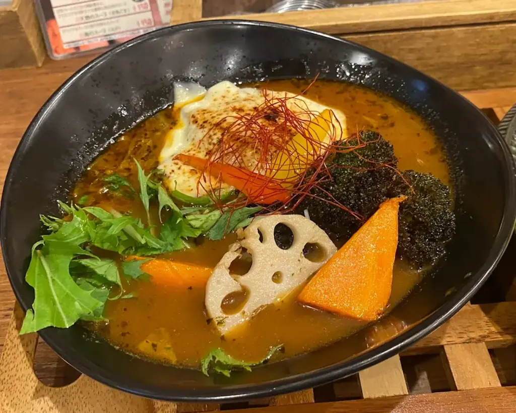 A vibrant bowl of Japanese curry soup with a slice of grilled cheese, lotus root, carrot, broccoli, and leafy greens. Garnished with red pepper threads.