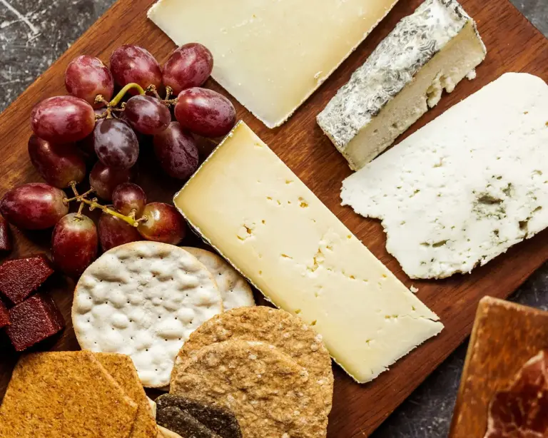 An assortment of cheeses, crackers, and grapes on a wooden board. The scene conveys a rustic, inviting tone with a variety of textures and colors.