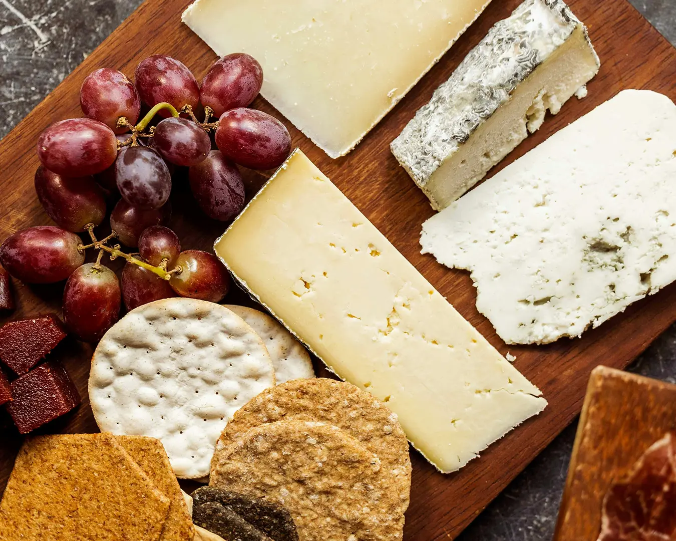 An assortment of cheeses, crackers, and grapes on a wooden board. The scene conveys a rustic, inviting tone with a variety of textures and colors.