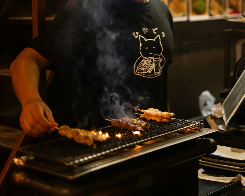An authentic scene of a chef in a graphic t-shirt cooking various yakitori skewers over a smoking hot charcoal grill, capturing the flame flare-ups and traditional cooking methods at a Singapore izakaya.