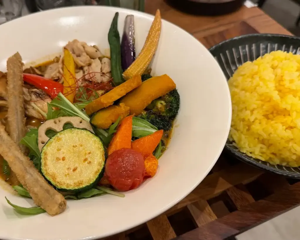 A vibrant bowl of Japanese curry soup with colorful vegetables like zucchini, carrot, eggplant, and okra, paired with a plate of turmeric rice.