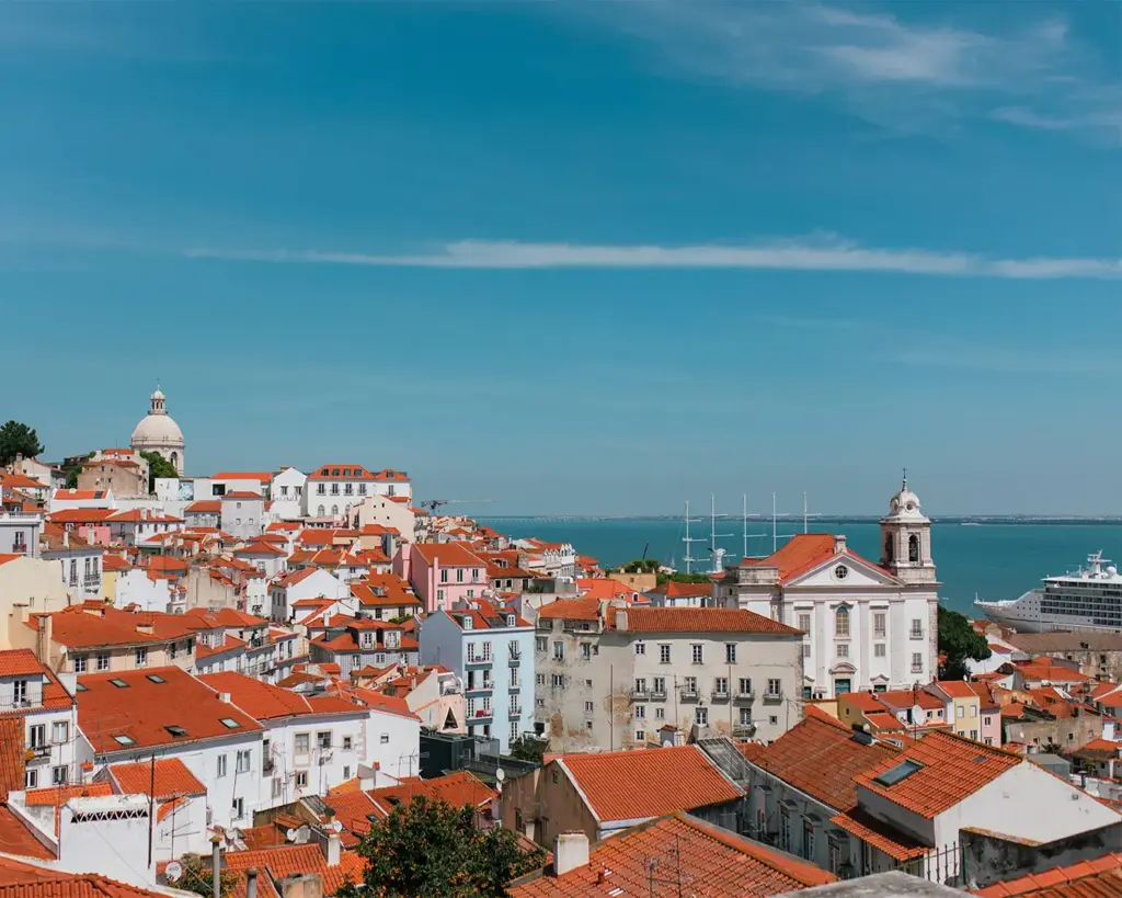 Scenic view of Lisbon’s Alfama district with red-tiled rooftops, historic buildings, and a church under a clear blue sky, overlooking the Tagus River.