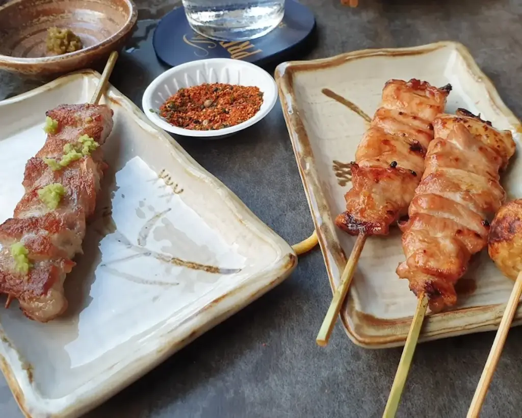 Alt text: Two ceramic plates with skewers of grilled meat. One skewer is topped with green paste. A dish of red seasoning and a glass are in the background.