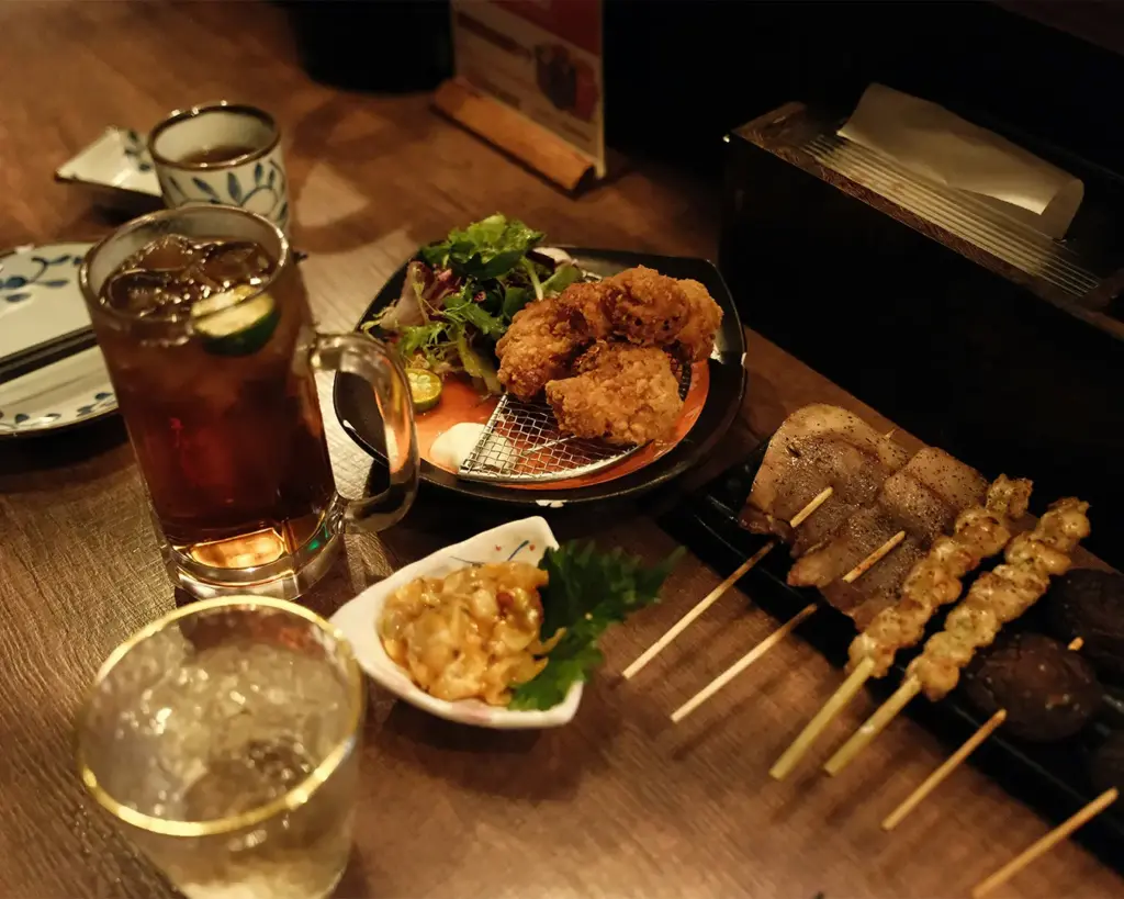 A full Japanese izakaya dining spread on a wooden table featuring assorted grilled yakitori skewers, crispy chicken karaage with a side salad, small dipping side dishes, an iced beverage, and a glass of sake.