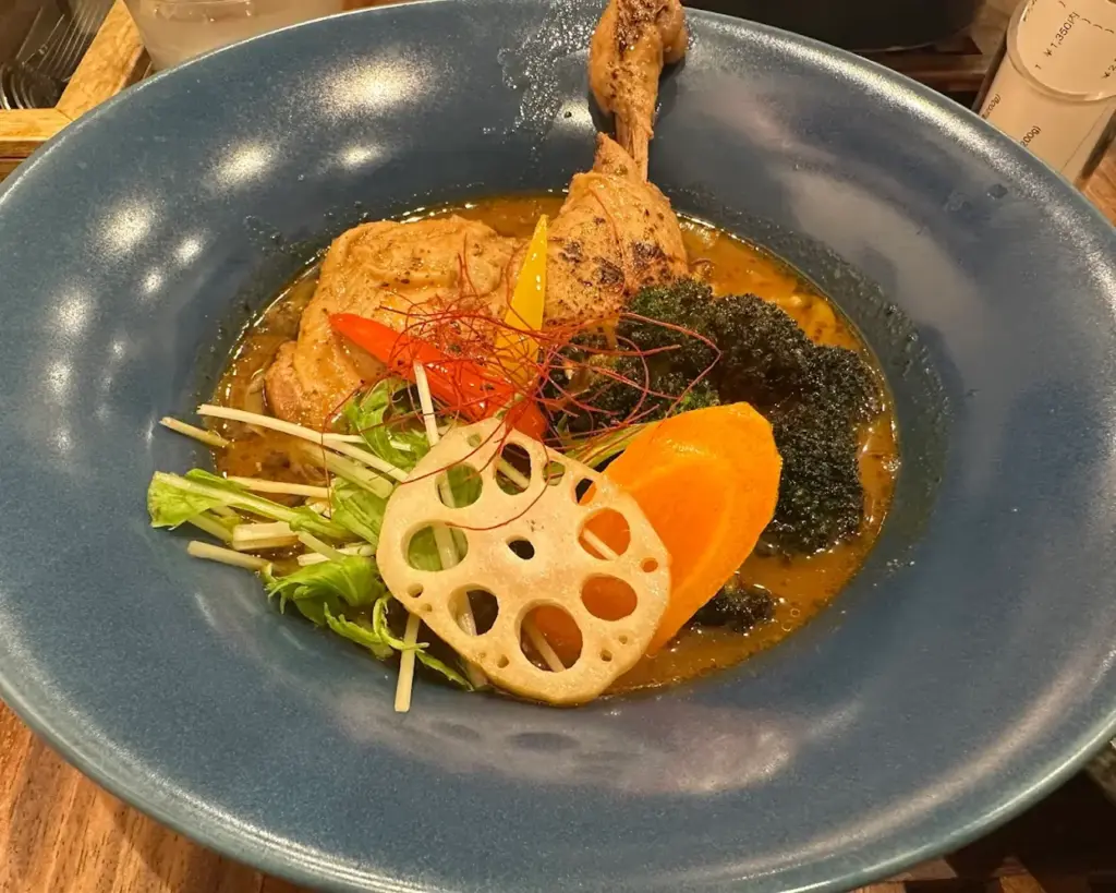 A vibrant dish in a blue bowl featuring a roasted chicken leg, colorful vegetables, lotus root slice, carrot, and greens in a rich sauce.