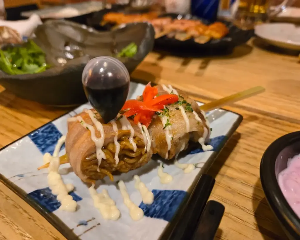 Grilled noodle skewer wrapped in bacon with drizzled mayo, topped with red garnish and herbs, served on a decorative plate with soy sauce.