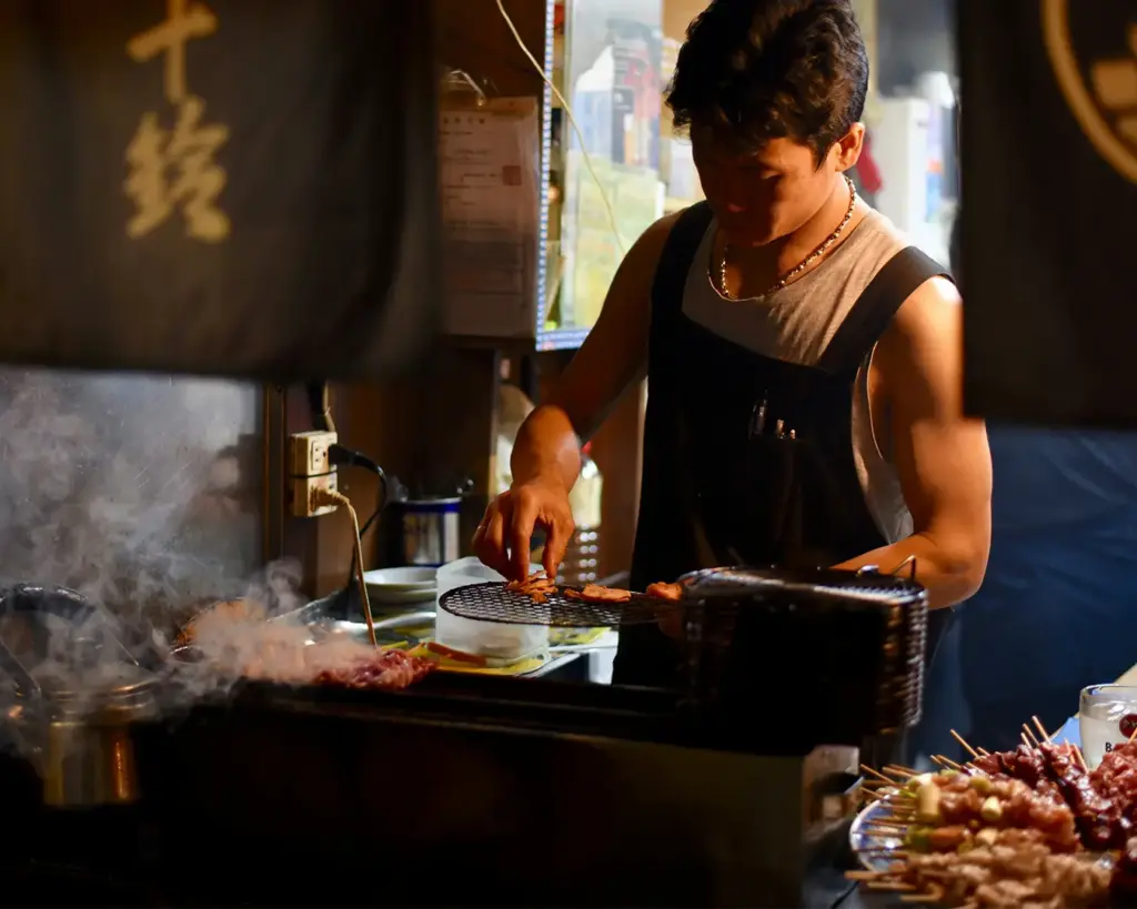 A focused Japanese yakitori chef carefully arranging and grilling raw chicken meat skewers on a traditional charcoal grill station filled with smoke and warm ambient lighting inside a bustling restaurant.