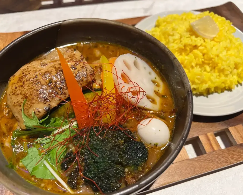 A vibrant bowl of curry with grilled chicken, lotus root, broccoli, and sliced peppers, garnished with herbs and red strands, served with yellow rice.