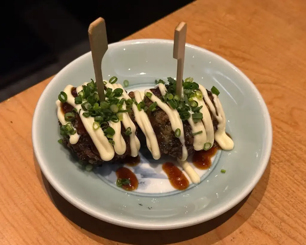 Two takoyaki balls on a white plate, topped with drizzled mayonnaise, chopped green onions, and sauce, secured with wooden picks, on a wooden table.