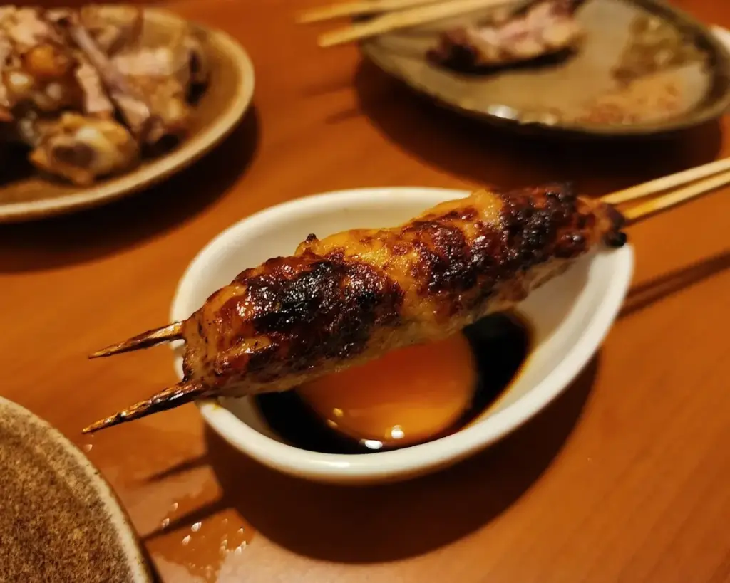 Grilled skewered meat placed over a dish of soy sauce and a raw egg yolk on a wooden table. Plates with more food are in the blurred background.